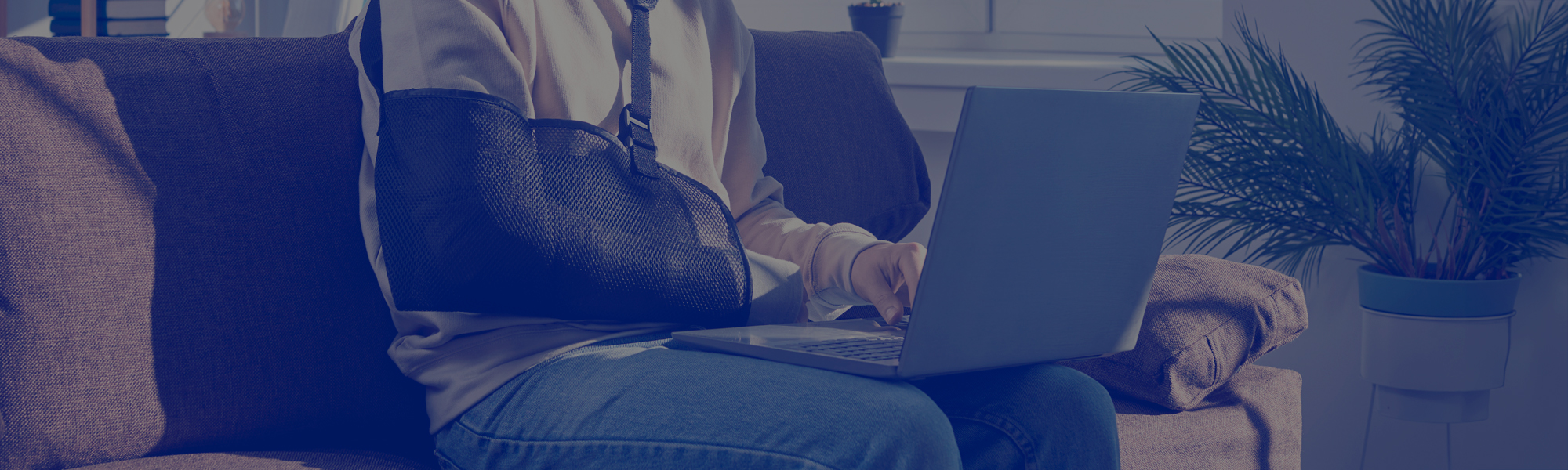 Man with a broken arm in an arm sling sitting on the sofa and working on his laptop computer